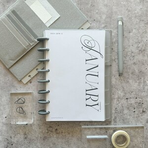 May include: A gray planner with a white cover and the text "2023 | OTR |" and "JANUARY CHAPTER I" in black. The planner has a silver ring binding and is open to the January page. The planner is on a gray surface with other office supplies.