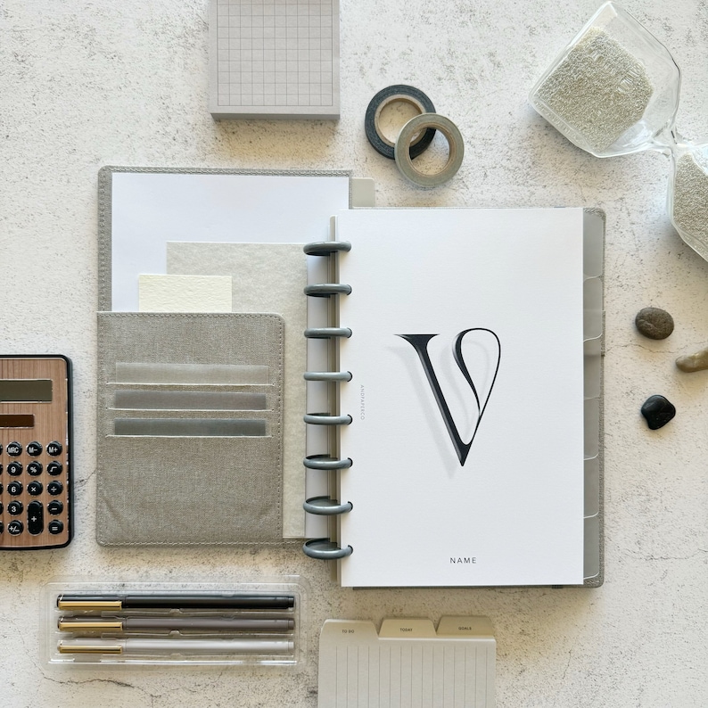 May include: A gray and white planner with a black letter V on the cover. The planner has a ring binding system and a pocket on the front cover. The planner is on a white surface with other office supplies.