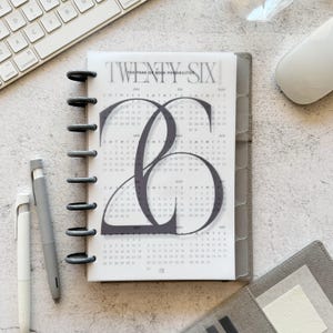 May include: A gray planner with the text "TWENTY SIX" and a calendar design. The planner is open, revealing monthly calendars. Two pens and a keyboard are visible in the background. The planner is bound with black rings.