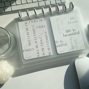 May include: A clear, binder-style planner with silver rings, holding two white cards. One card reads "I WILL BE SUCCESSFUL" and the other says "life is beautiful." The planner and cards are on a white surface.