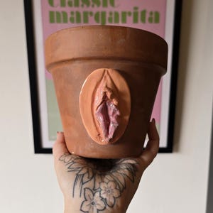 Könnte beinhalten: Ein Terrakotta-Pflanztopf mit einer skulpturalen, dreidimensionalen anatomischen Vulva-Darstellung in Pfirsich- und Rosatönen auf der Vorderseite. Der Topf hat einen breiten, kreisförmigen Rand und eine konische Basis mit einer texturierten Oberfläche.
