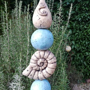 Könnte beinhalten: Eine gestapelte Gartenskulptur mit einer tropfenförmigen Form mit einem Spiraldesign, einer hellblauen Kugel, einer Muschel und einer weiteren hellblauen Kugel. Die Skulptur steht vor einem Hintergrund aus grünem Laub.
