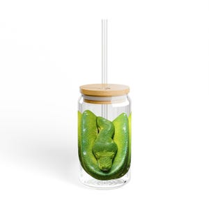 May include: A clear glass tumbler with a bamboo lid and a glass straw. The tumbler features a vibrant green snake design. The snake is coiled inside the glass, with its head facing forward. The tumbler is designed for cold beverages.