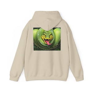 Könnte beinhalten: Beige Kapuzenpullover mit einer Illustration einer leuchtend grünen Schlange auf dem Rücken. Die Schlange hat gelbe Augen und eine rote Zunge. Das Design ist vor grünem Laub platziert und erzeugt einen auffälligen Kontrast.