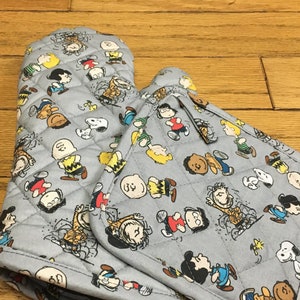 May include: A pair of gray oven mitts with a quilted design featuring a pattern of Peanuts characters. The characters are in various poses and include Snoopy, Charlie Brown, Lucy, and Linus.