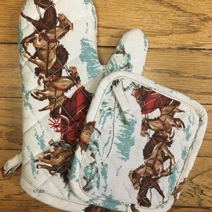 May include: A set of two white quilted oven mitts with a blue and white pattern of horses. The mitts are quilted and have a loop for hanging.