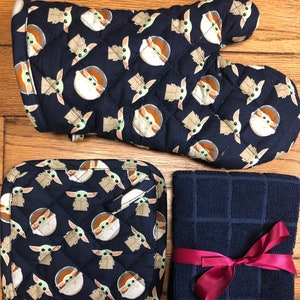 May include: A set of kitchen accessories featuring a blue and green pattern of a popular character from the Star Wars franchise. The set includes a pot holder, oven mitt, and a tea towel with a red ribbon.