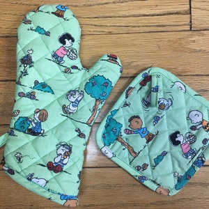 May include: A set of two green quilted oven mitts with a Peanuts Easter theme. The mitts feature characters from the Peanuts comic strip, including Snoopy, Charlie Brown, and Lucy, holding Easter eggs. The mitts are perfect for protecting your hands while cooking or baking.
