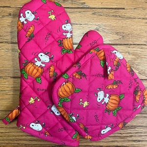 May include: A quilted pink oven mitt and pot holder set. The set features a repeating pattern of Snoopy, pumpkins, and the word "Boo!". The oven mitt has a loop for hanging. The pot holder is square.