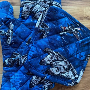 May include: A pair of blue quilted oven mitts with a Star Wars spaceship pattern. The mitts are made of a soft, quilted fabric and feature a variety of spaceships, including X-wings, TIE fighters, and the Millennium Falcon.