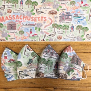 May include: Four fabric face masks with a white background and a colorful pattern of Massachusetts landmarks and text. The masks are made of cotton and have a blue and white trim. The text on the masks includes "Massachusetts", "Martha's Vineyard", "The Berkshires", "Minute Man National Historic Park", "Faneuil Hall", "Mayflower", "Cape Cod", "Nantucket", and "Black-capped Chickadee".