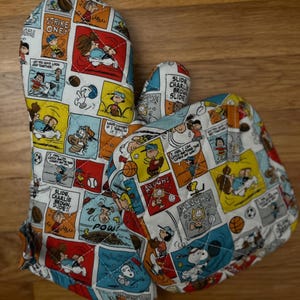 May include: A pair of quilted oven mitts featuring a comic strip design with characters and speech bubbles. The mitts are primarily white with colorful panels, including red, yellow, blue, and brown. Text includes "STRIKE ONE!" and "SLIDE CHARLIE BROWN, SLIDE!!".