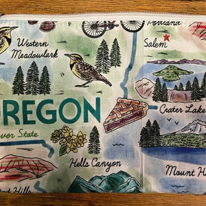 May include: A white zippered pouch with a map of Oregon. The map features various landmarks, including Crater Lake, Mount Hood, Painted Hills, and Hells Canyon. The pouch also features the text "Oregon, Beaver State" and a Western Meadowlark.