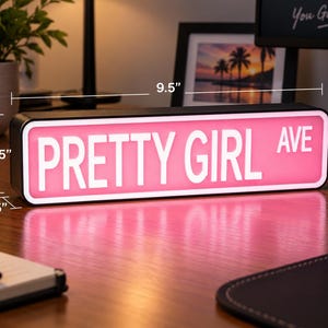 May include: A rectangular LED sign with a pink background and white text reading PRETTY GIRL AVE. The sign measures 9.5 inches in length, 3.25 inches in height, and 1.25 inches in depth, featuring a black frame and a bright, illuminated display.