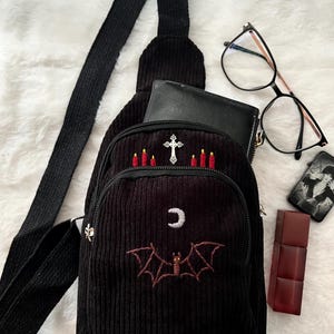 May include: Black corduroy sling bag with embroidered bat and crescent moon design. Features a cross and candle embroidery on the top pocket. Includes a black strap, zipper closure, and a small black wallet.