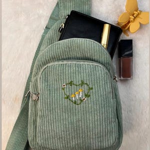 以下が含まれることがあります： キノコと花が刺繍されたハート型のデザインが施された、緑色のコーデュロイ製のクロスボディバッグ。バッグはジッパーで閉じられ、調節可能な長いストラップが付いています。