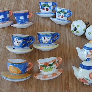May include: A collection of miniature ceramic tea sets, including teacups with saucers, teapots, and decorative knobs. The teacups feature various designs, such as cherries, cats, and orange slices. The teapots are decorated with flowers and apples.