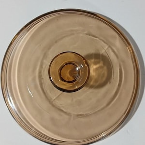 May include: A circular, amber-colored glass lid featuring a smooth, translucent surface and a centered, raised knob handle. The lid has a slightly elevated rim and a consistent, warm brown tint throughout the entire glass structure.
