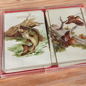 May include: Vintage playing cards in a clear plastic case. The cards feature illustrations of a largemouth bass and birds. The artwork is detailed with natural colors, set against a backdrop of a river and trees.