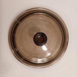 May include: A round, brown glass lid with a dark brown knob in the center. The lid has a slightly raised rim and is designed for cookware. The glass is transparent, allowing visibility of the contents.