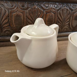 May include: A small, off-white ceramic teapot with a matching lid. The teapot has a rounded body, a curved spout, and a small handle. The lid has a rounded knob for easy removal. The teapot is sitting on a wooden surface.