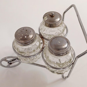 May include: A vintage glass salt and pepper set with a metal carrier. The clear glass shakers have a textured pattern and silver-colored lids. The carrier has a handle and a wheel-like design on the side, creating a unique kitchen accessory.