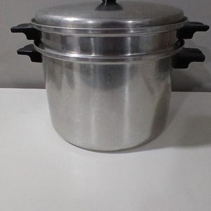 May include: A silver-colored aluminum steamer pot with a matching lid. The pot features two black plastic side handles, and the lid includes a black plastic knob handle. The steamer insert is nested within the main pot, creating a two-tier design.