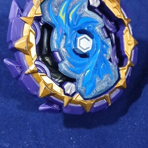 May include: A circular spinning top toy featuring a purple outer ring with gold-colored triangular spikes. The center displays a blue translucent plastic insert with a stylized dragon graphic and a central hexagonal silver-colored bolt component.