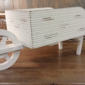 May include: A small, white wooden wheelbarrow with a distressed finish. The wheelbarrow has a single wheel, a rectangular bed, and two handles. The wheel is round with spokes. The wheelbarrow is sitting on a wooden surface.