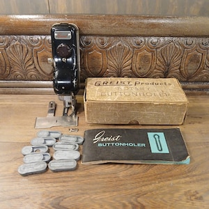 May include: A vintage black Greist rotary buttonholer sewing machine with a box and instruction manual. The box is tan with black text. The instruction manual is black with a teal rectangle. There are several gray buttonhole templates.
