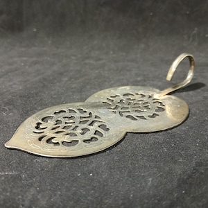 May include: A silver-toned metal tea strainer featuring a dual-lobed, leaf-like shape with intricate, openwork scroll patterns throughout the surface. The piece includes a curved handle extending from one end, designed for resting on a cup rim.