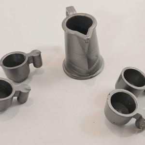 May include: A set of five miniature silver-colored plastic items consisting of one small pitcher with a handle and four matching small cups with handles. Each piece features a smooth, solid finish and a simple, functional design for play.