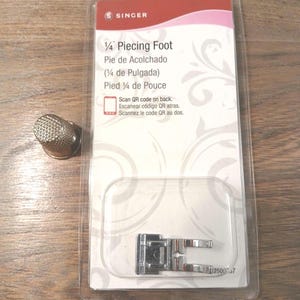 May include: A silver-colored metal thimble and a Singer 1/4 inch piecing foot for a sewing machine, packaged in a clear plastic blister pack with a white and red cardboard backing featuring the text 1/4 Piecing Foot and Scan QR code on back.