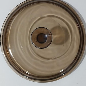 May include: A circular, translucent brown glass lid featuring a smooth, slightly domed surface and a centered, integrated knob handle. The lid has a reinforced outer rim and a polished finish, providing a clear view through the tinted material.