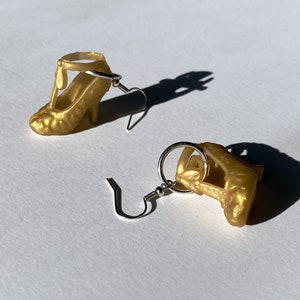 Könnte beinhalten: Ein Paar goldfarbene Ohrringe in Form von High Heels. Die Ohrringe sind Miniaturrepliken von High Heels, komplett mit Riemen und Absätzen. Die Ohrringe sind an silberfarbenen Haken und vor weißem Hintergrund.