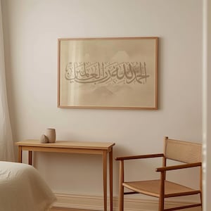 May include: A framed artwork featuring Arabic calligraphy in a light brown tone is displayed on a cream wall. The artwork is in a wooden frame. A wooden table and chair are positioned in front of the wall. A vase with two decorative stones is on the table.