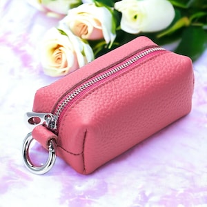 May include: A small, rectangular, pink leather pouch with a silver zipper and a silver ring attached. The pouch has a textured surface and a pink zipper. The background features white roses.