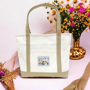 May include: A white canvas tote bag with beige straps and a beige bottom. The bag has a small pocket on the front with an embroidered cartoon image and the text "Days with friends should never end!"