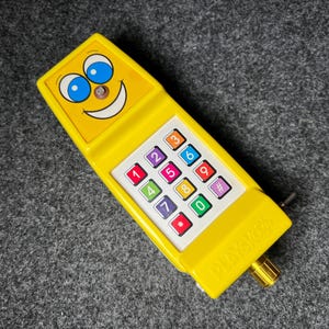May include: A vintage yellow toy phone with a smiling face and blue eyes. The phone has a colorful keypad with numbers and symbols. The word "PLAYSKOOL" is printed on the side. A classic toy for children.