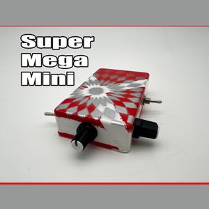May include: A rectangular effects pedal labeled Super Mega Mini with a red and silver geometric star pattern. The device features two black control knobs on the front edge and two metal toggle switches on the side panels of the unit.