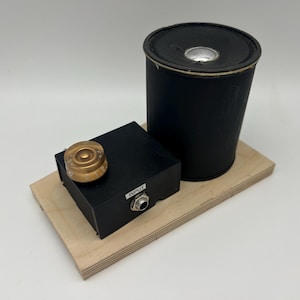 May include: A black speaker, a black box with a gold knob, and a wooden base. The black box has the word "INPUT" printed on it. The speaker is cylindrical and has a silver center.