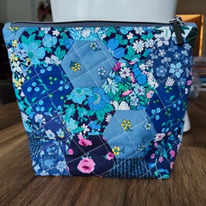 May include: A quilted cosmetic bag with a patchwork design. The bag features a variety of blue floral patterns and geometric shapes. The bag has a dark blue zip and is sitting on a wooden surface.