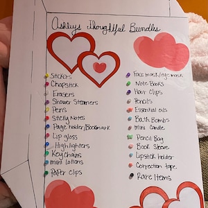 May include: A white paper with the text "Ashleys Thoughtful Bundles" and a list of items like stickers, lip gloss, and keychains. The paper is decorated with red hearts and colorful dots next to each item.