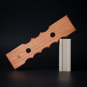 May include: A light-colored wooden wine bottle holder with a wavy edge and two circular holes. The word "STOIC" is printed on the wood. It is propped up by a white rectangular block against a black background.