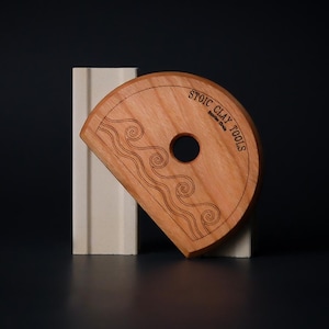 May include: A wooden clay tool with a semi-circular shape and a central hole. The tool is engraved with a wave pattern and the text "STOIC CLAY TOOLS". It is positioned next to a white rectangular block against a dark background.