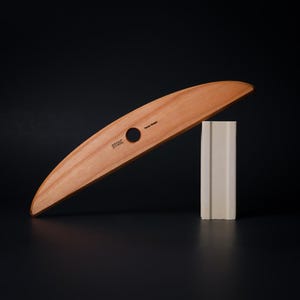 May include: A wooden pottery rib with the word "STOIC" and "Potter's Rib" printed on it. The rib is light brown and curved, with a circular hole. It rests on a white rectangular block against a black background.