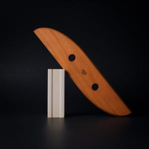 May include: A wooden pottery rib with two holes and the text "STOIC" is angled against a white rectangular clay form. The rib is light brown and the clay form is off-white, set against a black background.