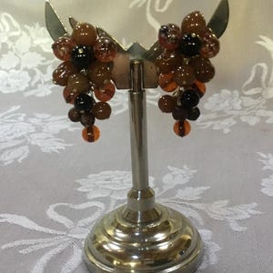 May include: A pair of vintage clip-on earrings featuring clusters of brown, amber, and black beads. The earrings are displayed on a silver-toned metal stand with a decorative base, set against a floral patterned background.