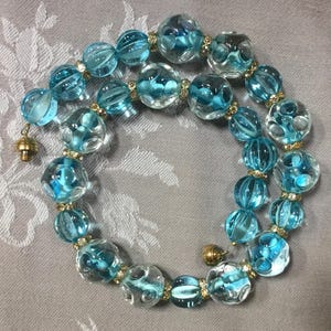 May include: A necklace with large, clear, and light blue glass beads. The beads are linked by small gold-coloured spacers. The necklace has a gold-coloured clasp. The beads have various shapes and internal designs.