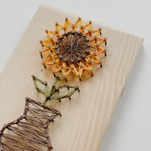 May include: A wooden board with a string art sunflower and vase. The sunflower is yellow and brown, and the vase is brown. The string art is created by wrapping string around nails hammered into the wood.